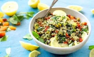 A tabbouleh bowl of couscous salad with vegetables and herbs sits on a light blue table. Fresh lemon wedges and mint leaves are scattered around, conveying freshness.