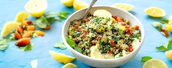 A tabbouleh bowl of couscous salad with vegetables and herbs sits on a light blue table. Fresh lemon wedges and mint leaves are scattered around, conveying freshness.